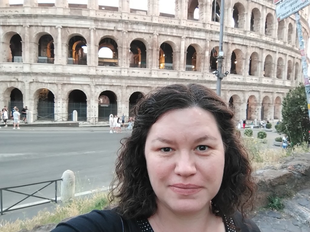 A picture of Phoebe Garrett, a woman with brown hair standing in front of the Colosseum in Rome